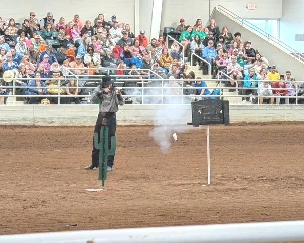 Someone standing on a dirt arena, possibly at a rodeo, with a shooting device producing smoke near a cactus-shaped object, while a large audience watches from the bleachers.