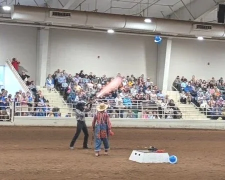 Cowboy and clown throwing a balloon on an indoor dirt arena with an audience seated in bleachers.