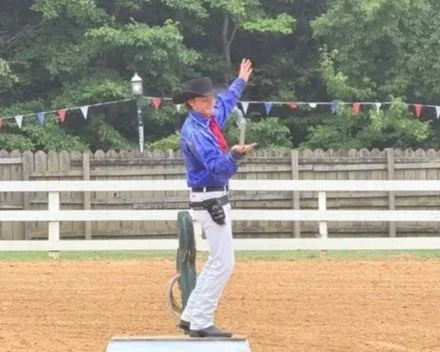 A person in country western attire, wearing a cowboy hat and sunglasses, holding a microphone, standing on a barrel in a rodeo or country event setting.