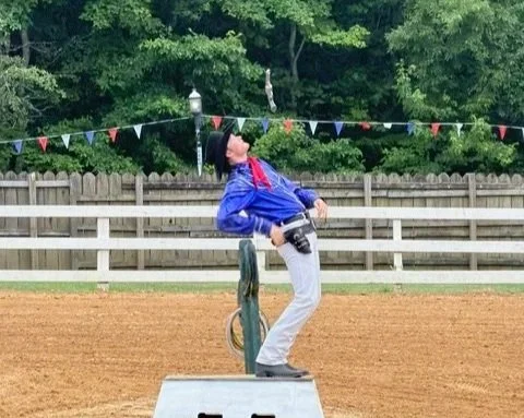 Person in blue jacket and gray pants standing on a platform near a baseball field, holding a camera, with trees and colorful bunting in the background.
