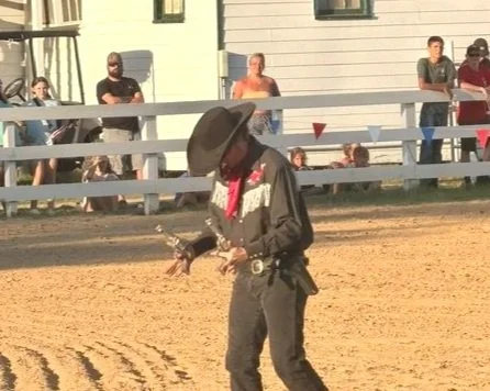 A cowboy wearing a hat and black outfit with a red scarf, standing on a dirt rodeo arena.