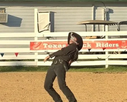 A person bending backwards on a dirt field, wearing a helmet and black clothing, with a sign that reads 'Texas Black Riders' in the background.