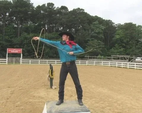 A person dressed in western attire, including a cowboy hat, blue jacket, and red scarf, stands on a raised platform in an outdoor arena, swinging a lasso.