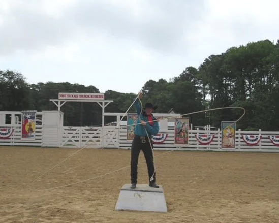 A cowboy performing a trick at the Texas Trick Ropers show, standing on a small platform and swinging a lasso in an outdoor arena with banners and trees in the background.