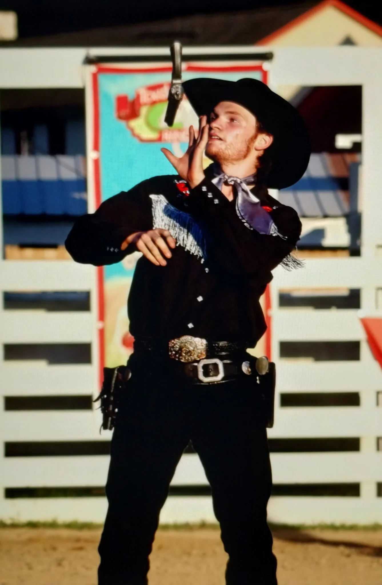 A man dressed as a cowboy with a black hat, black shirt, and western belt buckle, standing in front of a white fence at a rodeo or western themed event.