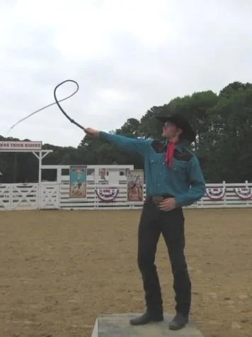 A cowboy in a blue shirt, black hat, and red neckerchief twirling a lasso in an outdoor rodeo arena with a decorated fence and a cloudy sky.