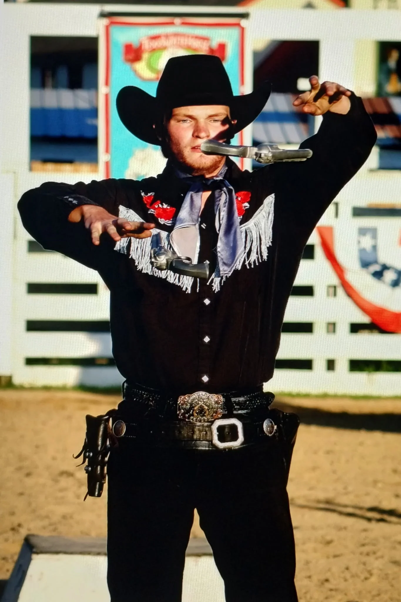 A man dressed as a cowboy, wearing a black hat and western attire, is performing a stunt with a gun balanced on his nose at a rodeo or western event outdoors.