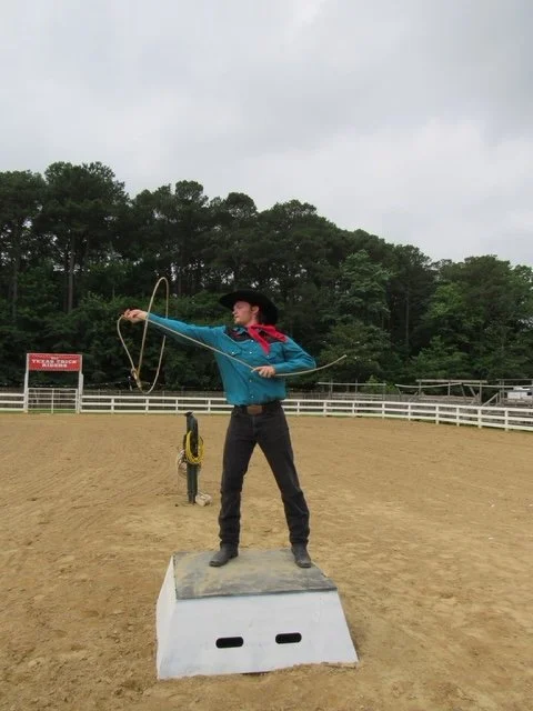 A person dressed as a cowboy in a blue shirt, red neckerchief, and black hat, standing on a platform and roping a target in an outdoor rodeo arena with trees in the background.