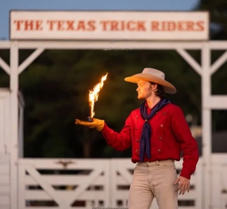 A person dressed as a cowboy at a Texas trick riding event, holding a flaming object in their right hand, wearing a large cowboy hat, red shirt, navy scarf, and beige pants, with a sign that reads 'The Texas Trick Riders' in the background.
