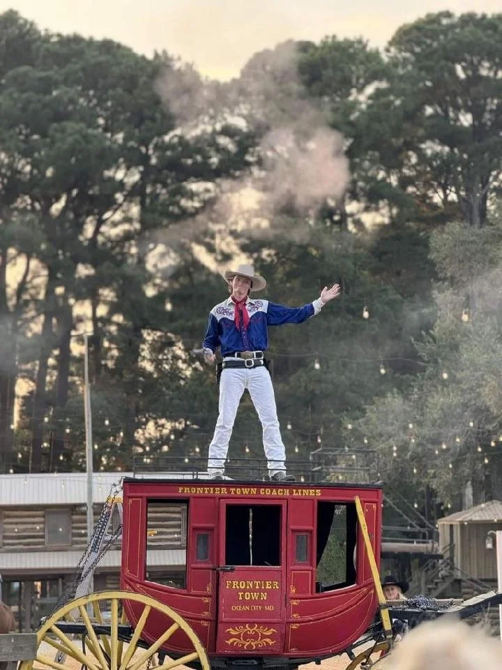A man dressed as a cowboy standing on top of a red wagon with the words 'Frontier Town' printed on the side, with trees and buildings in the background.