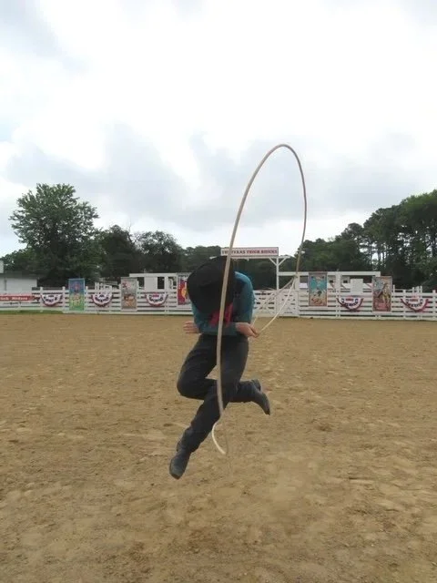 Person jumping with a lasso in an empty rodeo arena with a Texas flag-style banner backdrop.