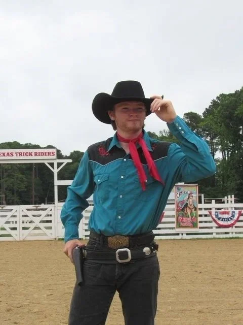 Man dressed as a cowboy at a rodeo or fairground, wearing a black cowboy hat, blue western shirt, red neckerchief, and belt with a large buckle, with a firearm in his pocket, tipping his hat.