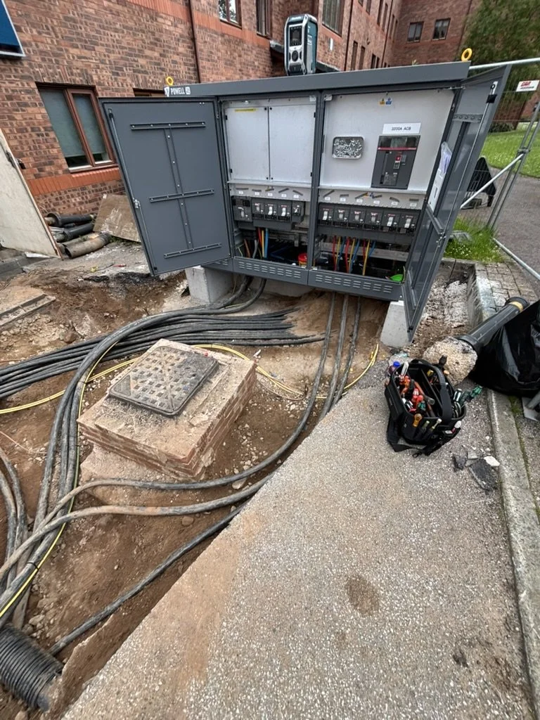 Open electrical control panel with various circuit breakers and wiring, outdoors on a construction site with tools nearby.