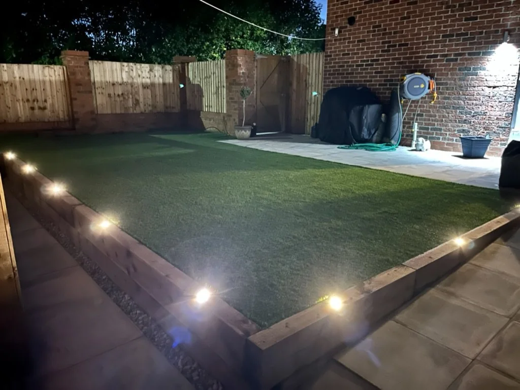 Backyard patio with a small yard illuminated by outdoor lights along a brick border, and a wooden fence surrounding the yard. There is a section with artificial grass, a potted plant, and a brick wall with garden equipment and a water hose.