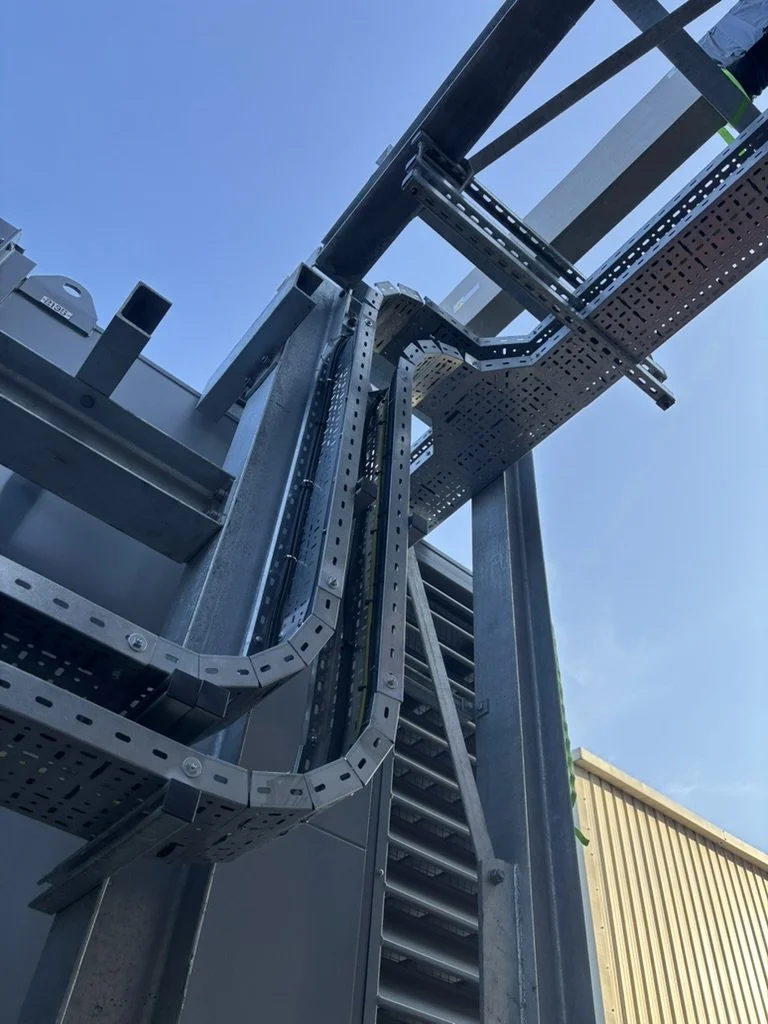 Close-up of industrial electrical or mechanical equipment with metal conduits, cable trays, and ladder supports against a blue sky.