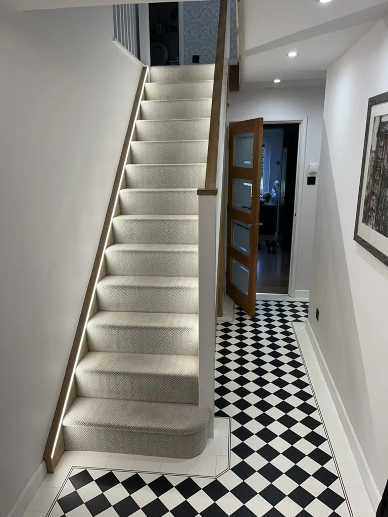 Interior view of a staircase with beige carpet leading to an upper floor in a home, adjacent to a hallway with black and white checkered tile flooring, a wooden door slightly open revealing a room, and framed artwork on the wall.