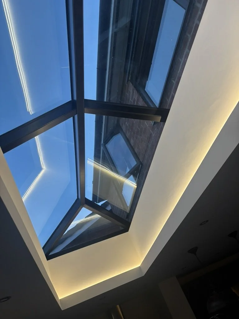 Inside view of a modern building showing a large rectangular glass skylight with multiple panes and a view of the blue sky outside, framed by interior ceiling with cove lighting.
