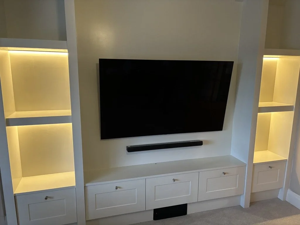 Flat-screen TV mounted on a white entertainment center with built-in shelves and drawers, illuminated with yellow lighting.