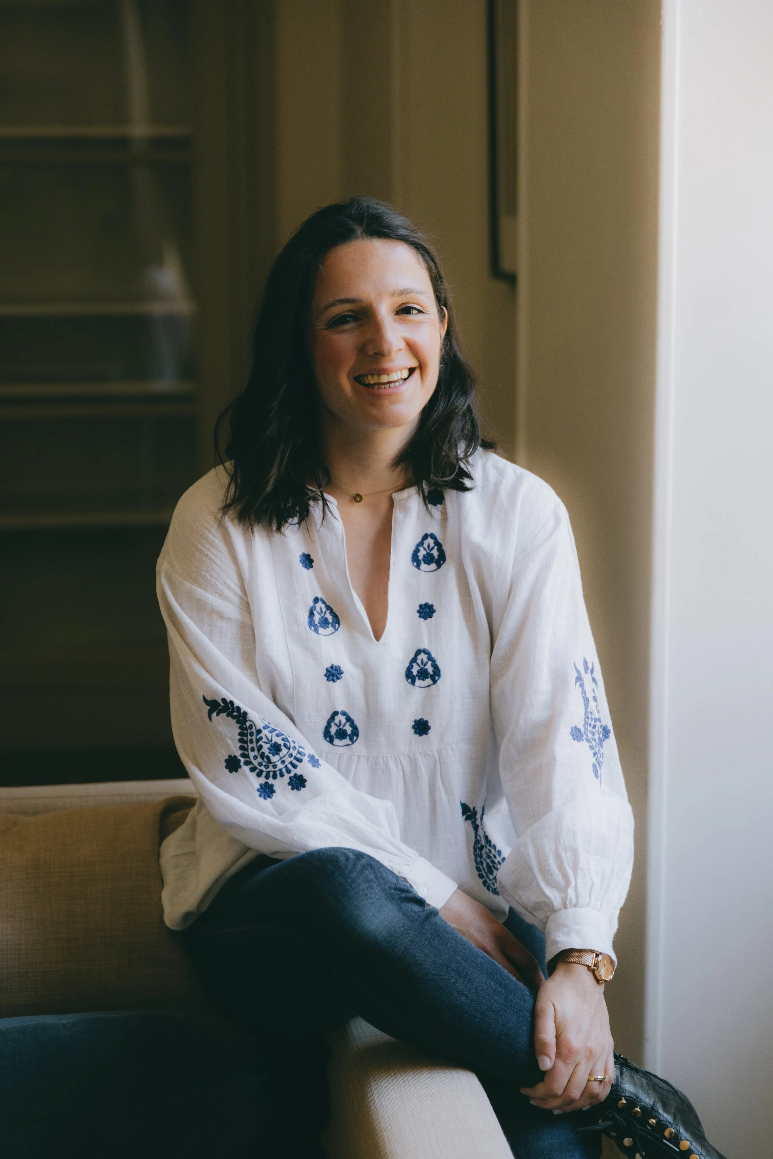 Jeune femme souriante avec des cheveux bruns, portant une blouse blanche à motifs bleus et assise dans une pièce lumineuse.