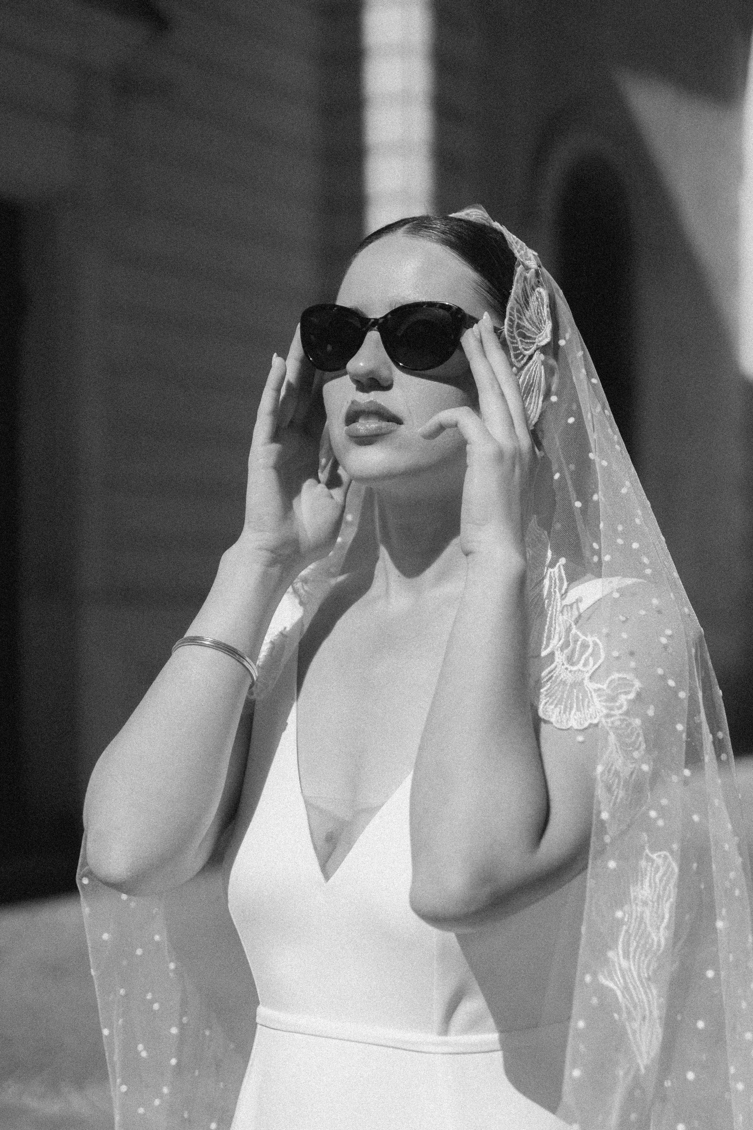 Une femme portant des lunettes de soleil foncées, un voile avec des détails en dentelle, et un maillot de bain blanc.
