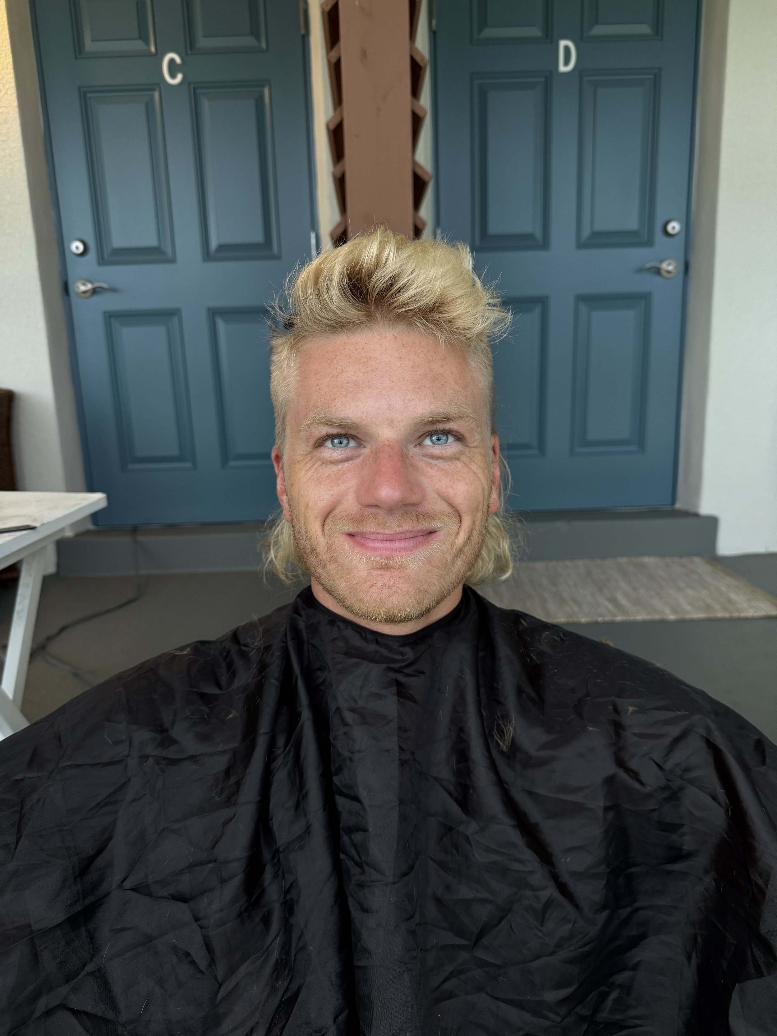 Daniel Joelo, singer and producer, smiling candidly during a haircut while wearing a black styling cape.