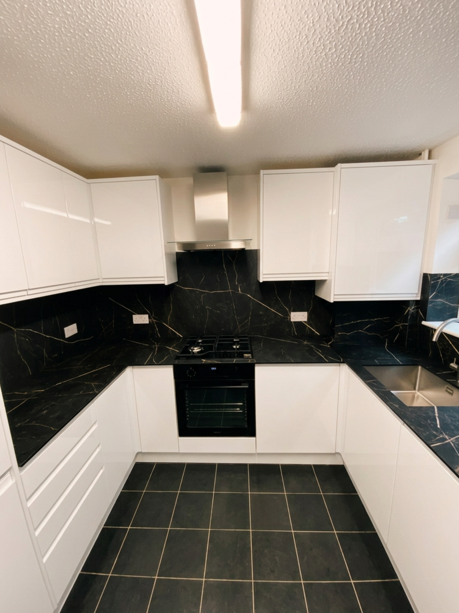 Professionally installed U-shaped kitchen featuring high-gloss white handleless cabinets, black marble-effect worktops and splashback with gold veining, built-in oven, gas hob, stainless steel extractor, and dark floor tiles. 
