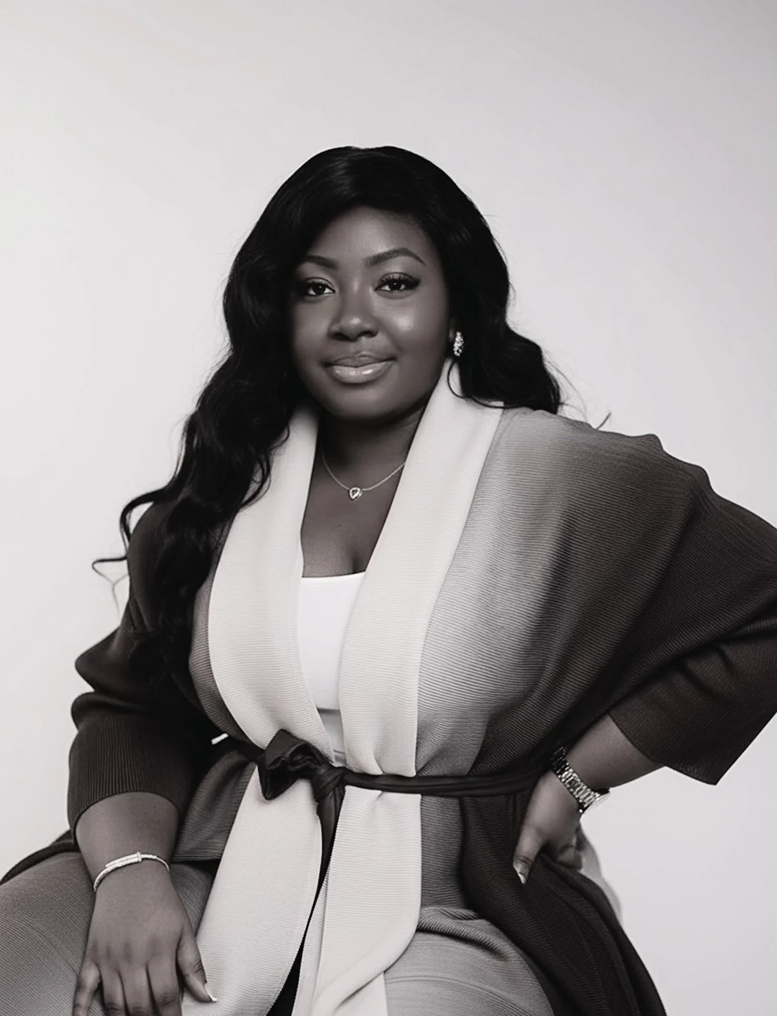 A Black woman with long, wavy hair wearing a black and white blazer, jewelry, and a watch, looking confidently at the camera against a plain background.