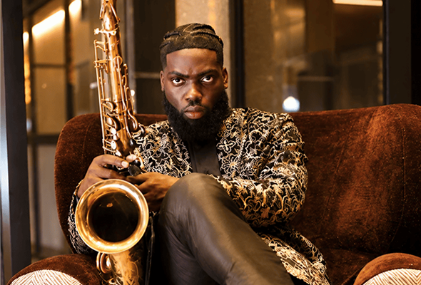 A man with a serious expression sitting on a brown couch, holding a saxophone, wearing a patterned jacket and black leather pants.