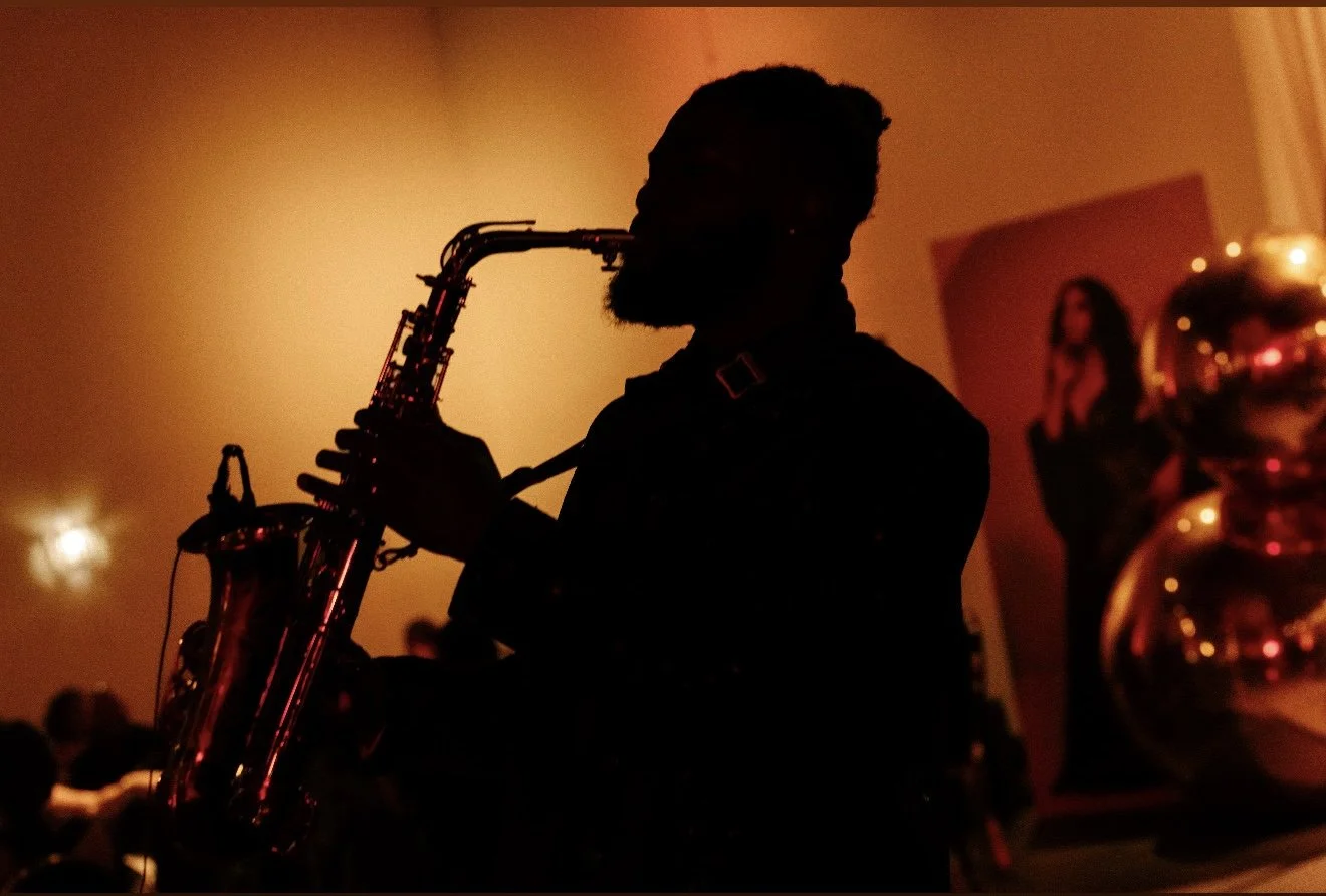 Silhouette of a man playing a saxophone in a dimly lit room with warm orange lighting, with a painting and decorative objects on the wall behind him.