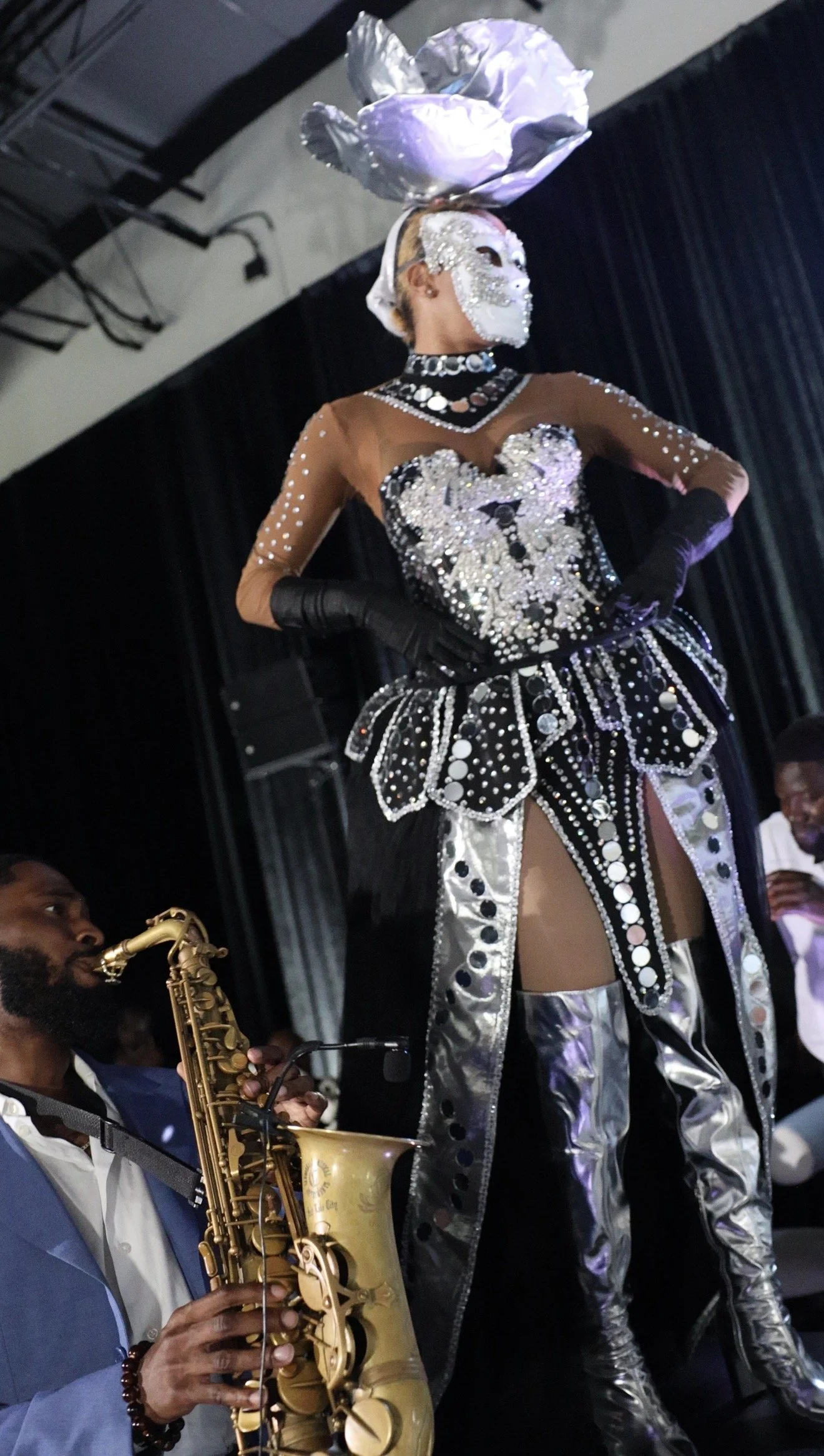 Performer in an elaborate silver and white costume with rhinestones, wearing a white mask and headpiece, on stage during a performance, accompanied by a musician playing a saxophone.