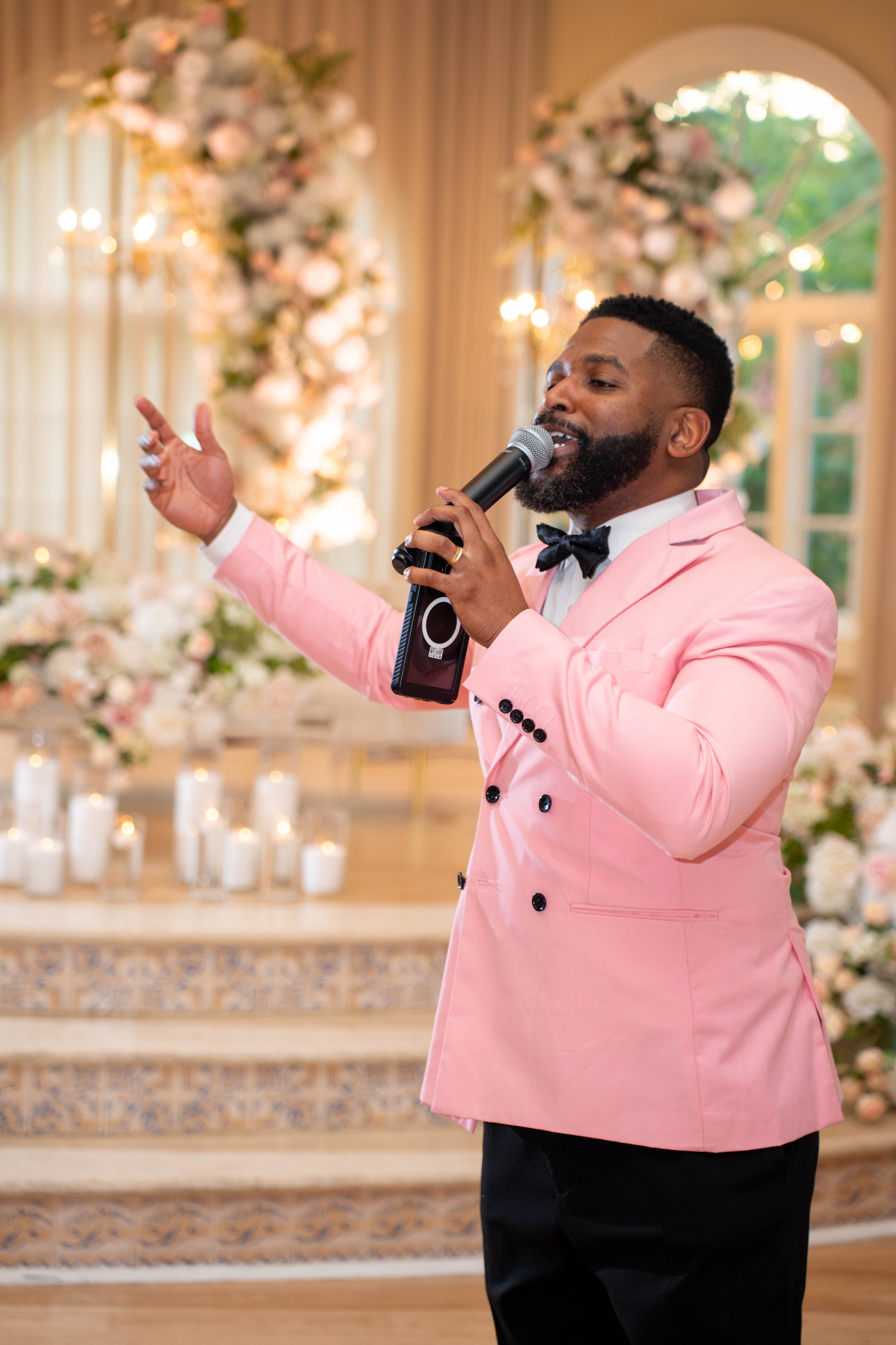 A man in a pink tuxedo jacket with a black bow tie, singing or speaking into a microphone at a decorated event venue with floral arrangements and candles in the background.