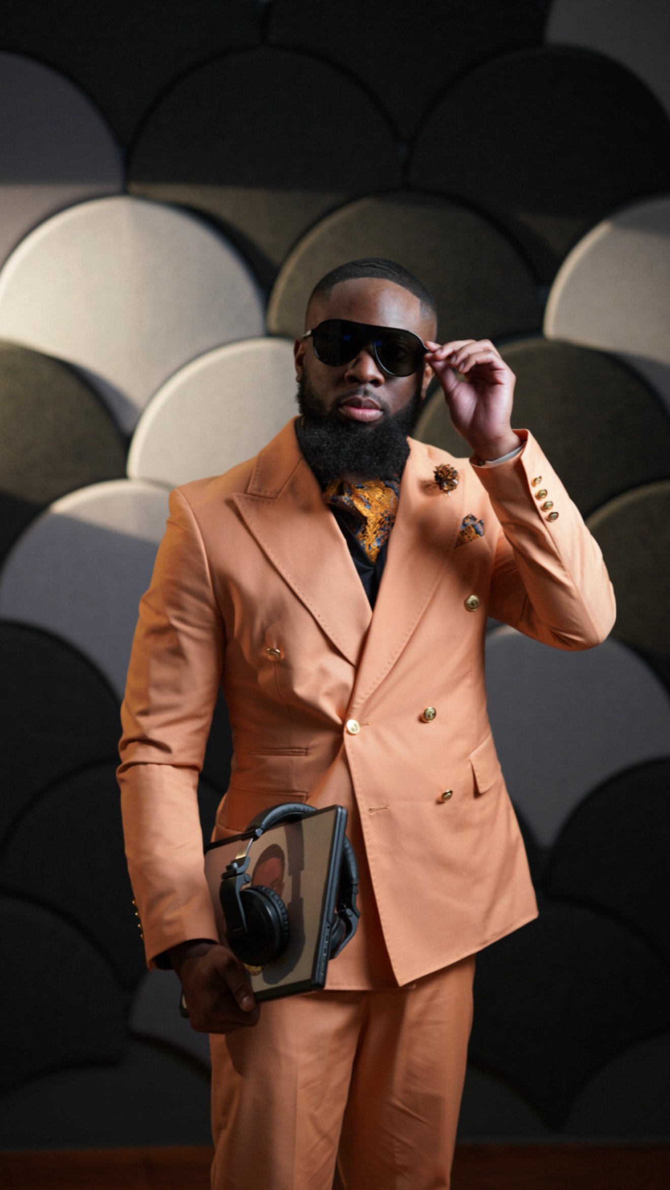 A man wearing a peach-colored suit, black sunglasses, and a patterned tie, holding a tablet with headphones on it, standing in front of a textured, circular wall background.