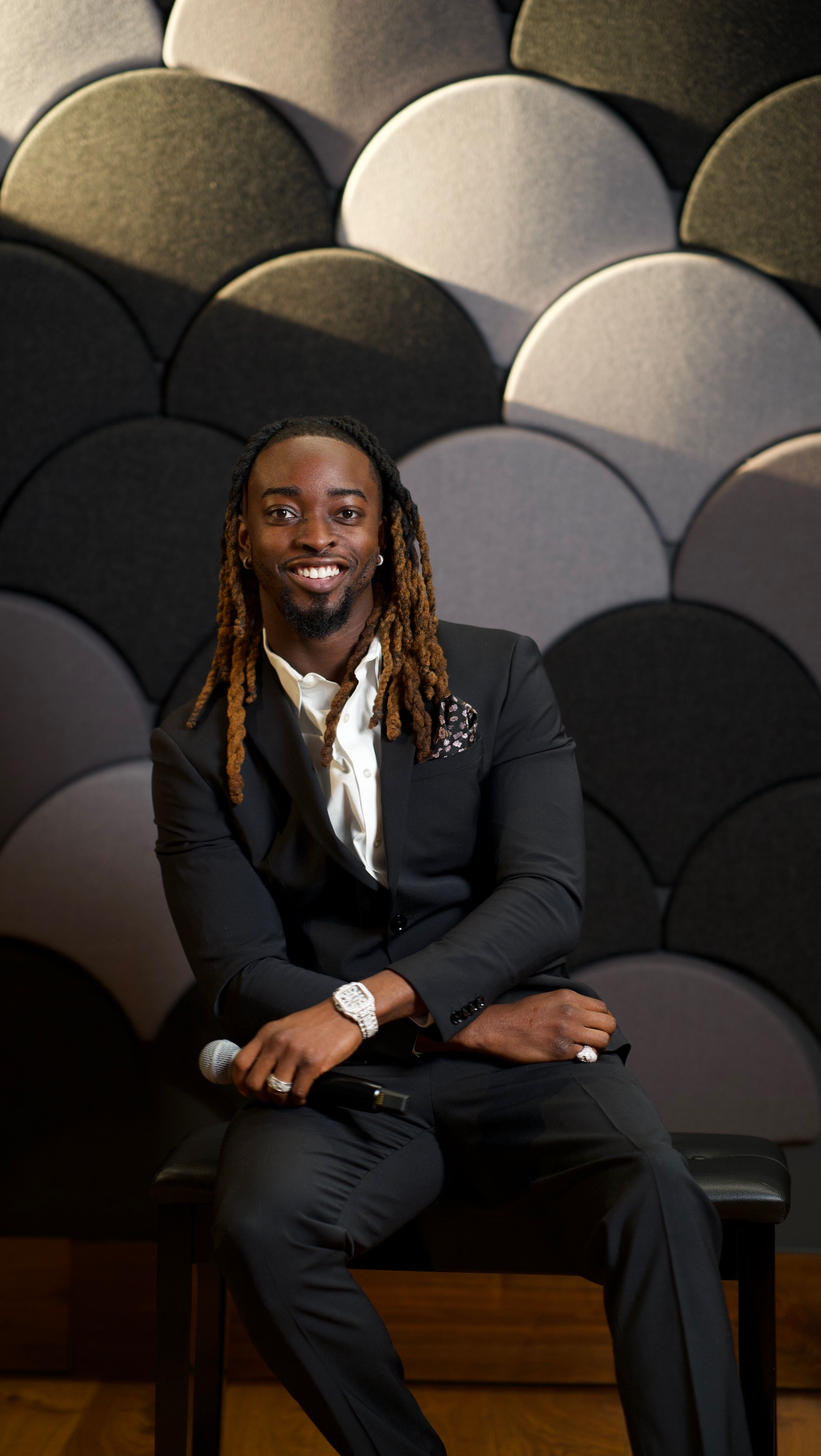 A man with long dreadlocks wearing a black suit, white shirt, and jewelry, sitting on a chair holding a microphone, smiling, with a patterned wall behind him.