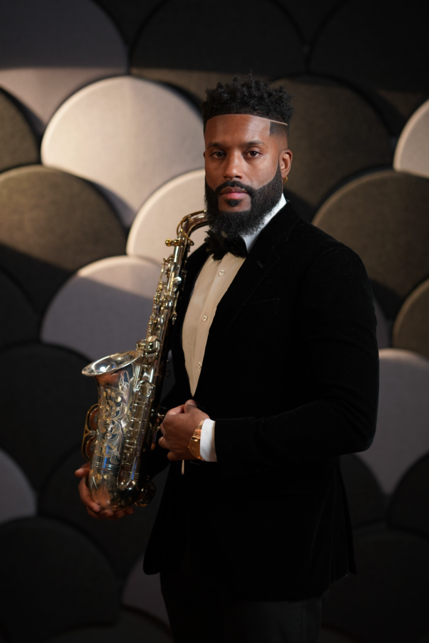A man dressed in a black tuxedo with a white shirt and black bow tie, holding a silver saxophone, standing in front of a background made of gray and black acoustic panels.