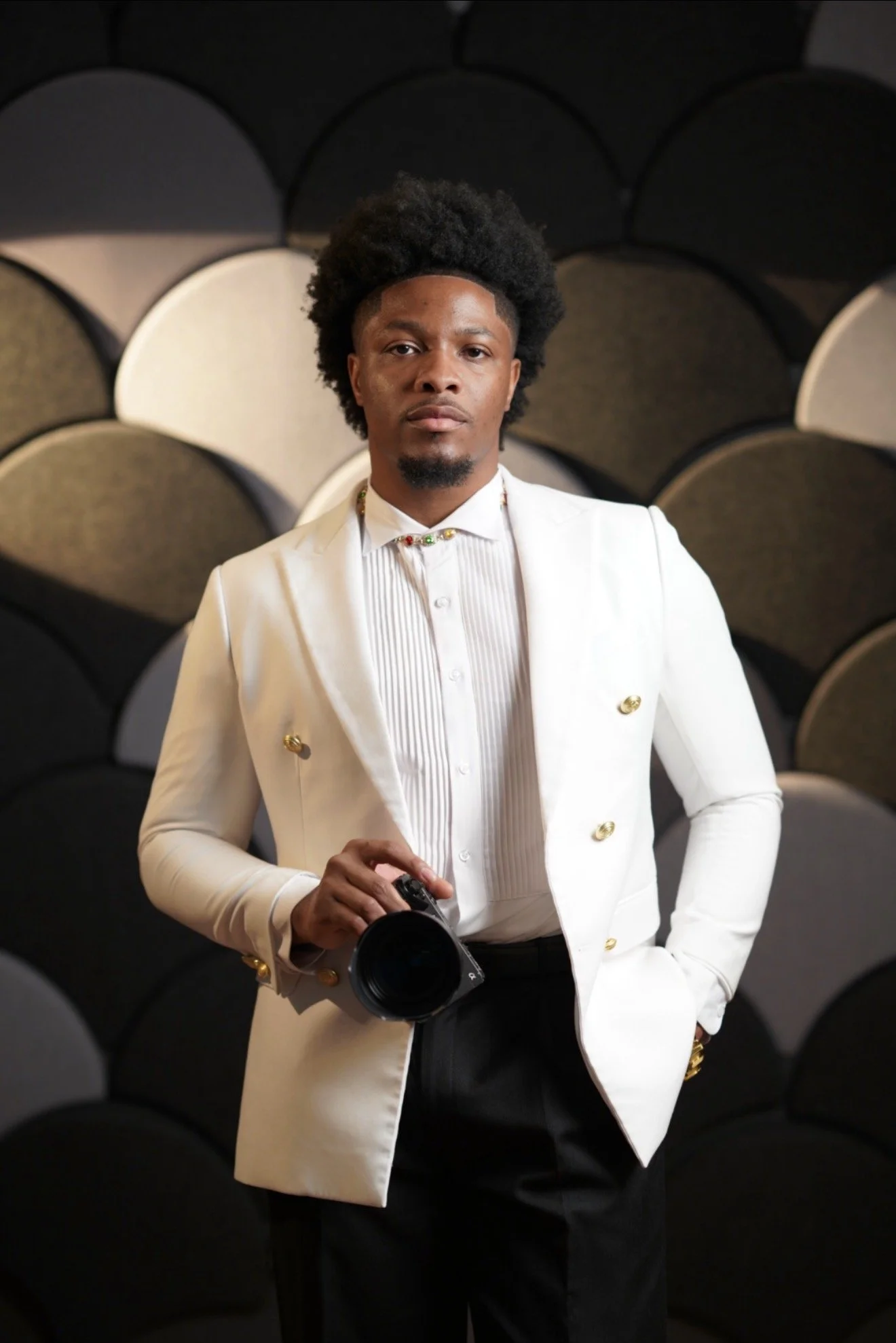 A man dressed in a white suit with gold buttons, holding a camera, standing in front of a background of circular acoustic panels.