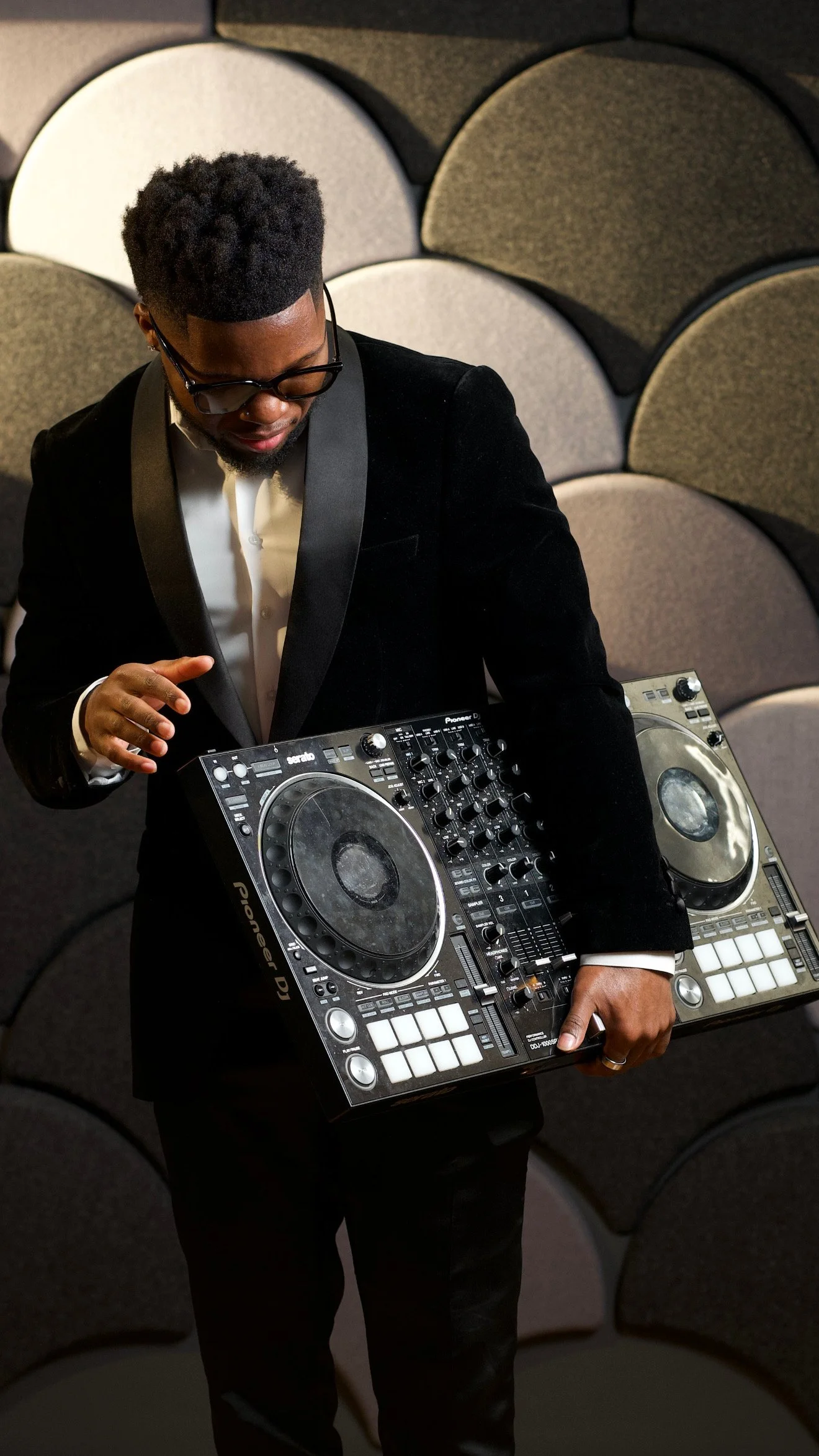 A man in a black tuxedo and glasses is holding a DJ controller and standing in front of a curved, textured wall.
