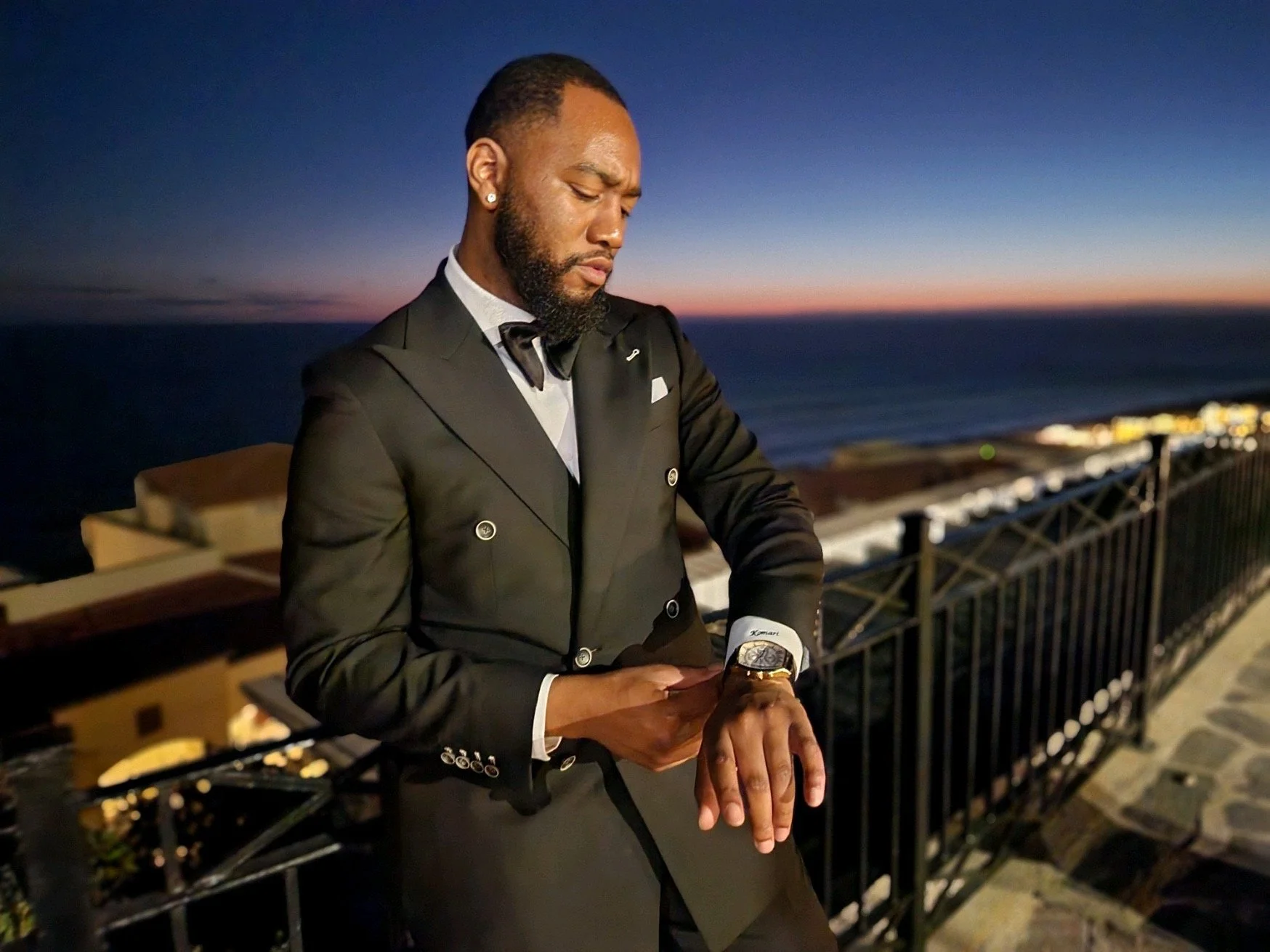 A man dressed in a black tuxedo with a bow tie standing outdoors on a balcony at sunset, looking at his wristwatch.