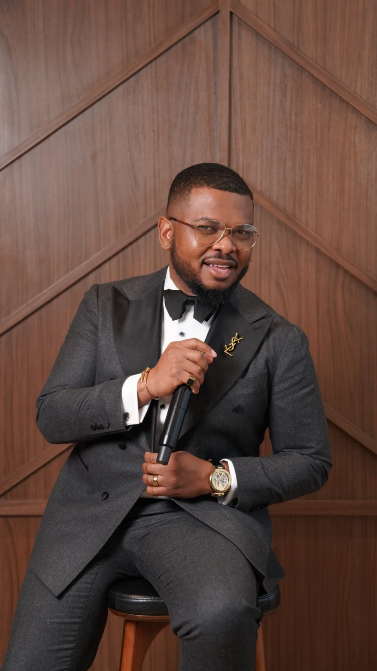 A man in a gray tuxedo with a bow tie, holding a microphone, sitting on a stool against a wooden-paneled wall, wearing glasses, a gold watch, and a YSL pin.