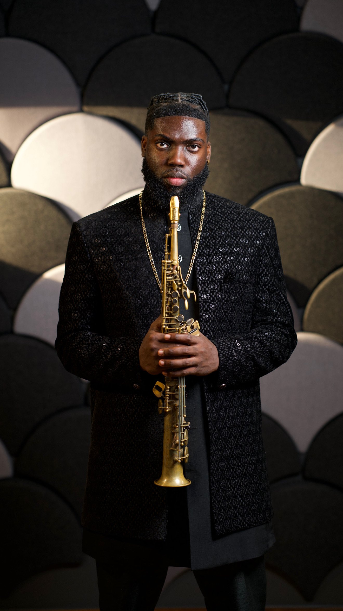 A man with a beard holding a golden saxophone in front of a background made of acoustic foam panels.