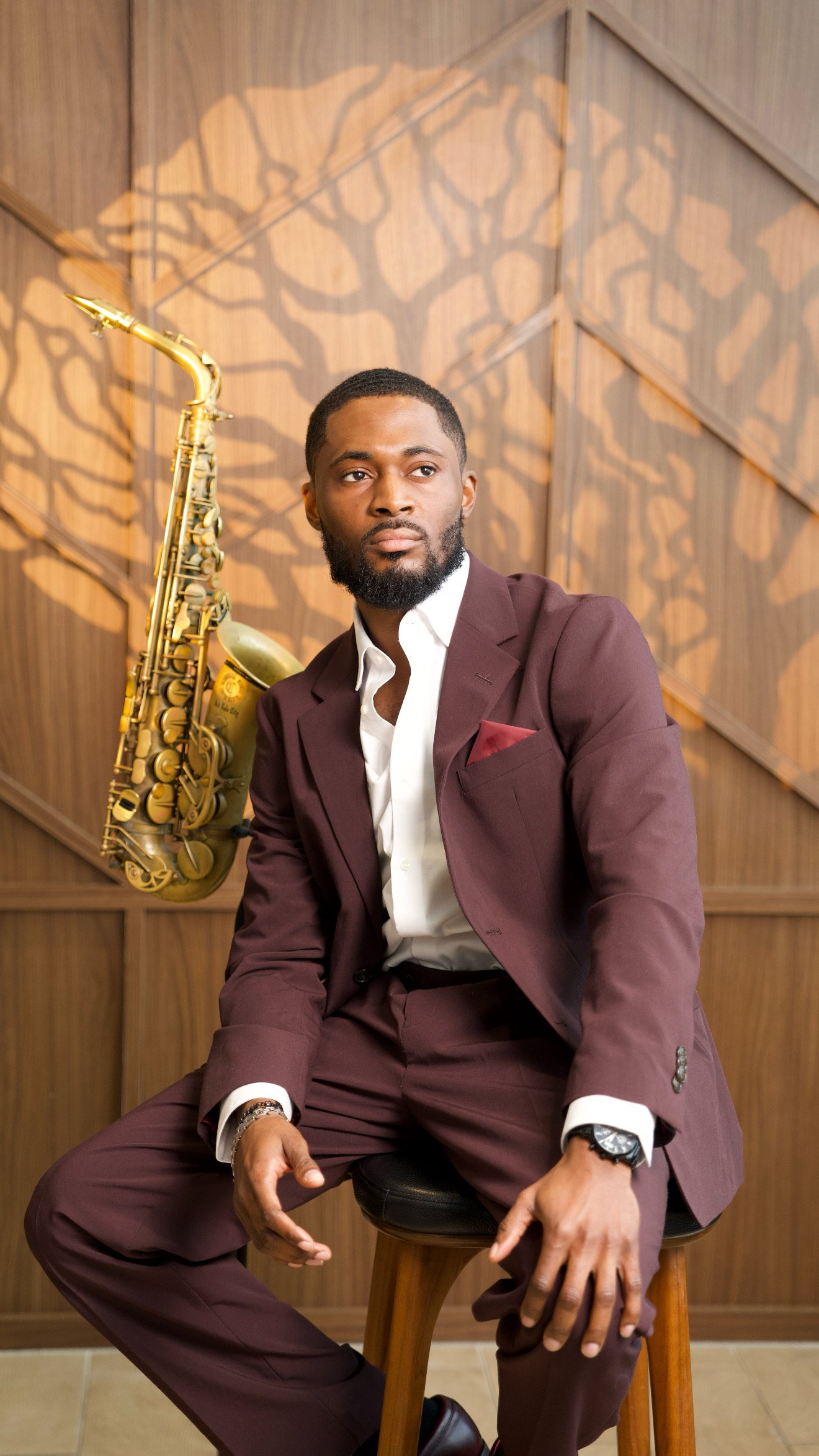 A man with a beard and short hair, dressed in a burgundy suit and white shirt, sitting on a wooden chair in front of a wooden wall with a tree-like shadow pattern, with a gold saxophone resting on his shoulder.