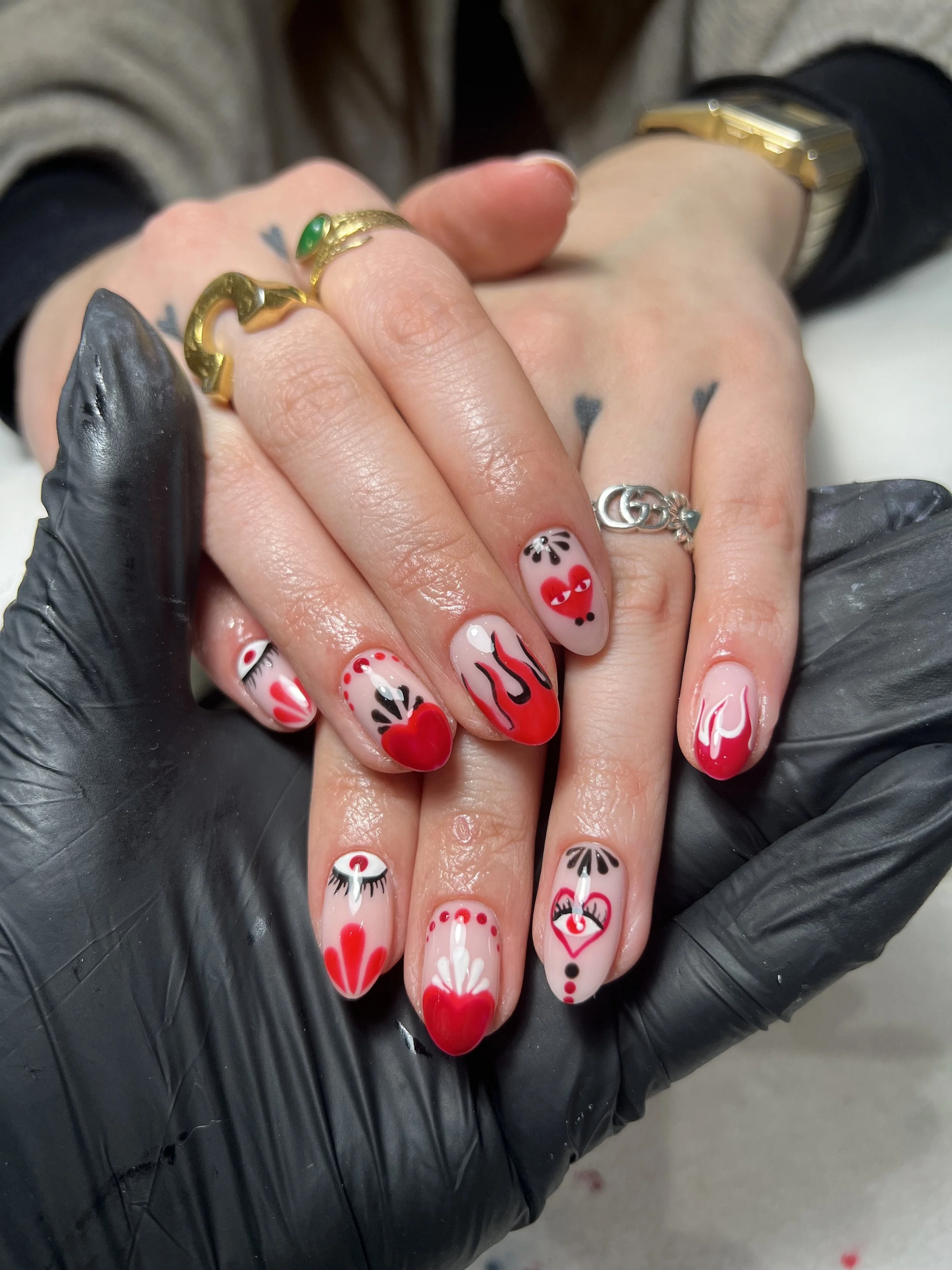 Close-up of a person's hand with decorated nails and jewelry, wearing black gloves and a watch, and holding another hand with a tattoo and jewelry.