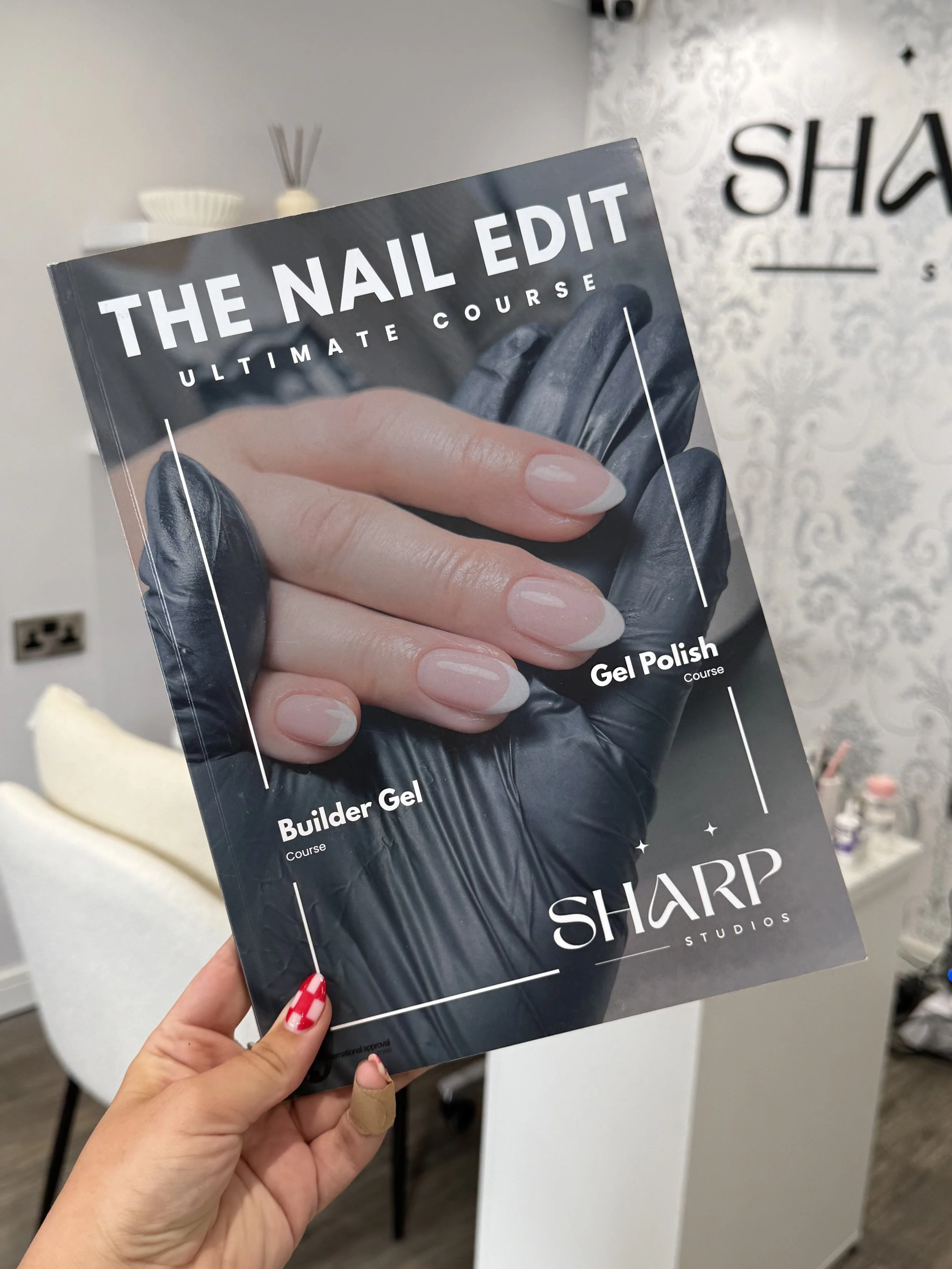 A person holding a beauty course brochure titled 'The Nail Edit: Ultimate Course' with an image of a hand with manicured nails and black gloves. In the background, there is a wall with a partial sign 'SHARP' and some salon products.