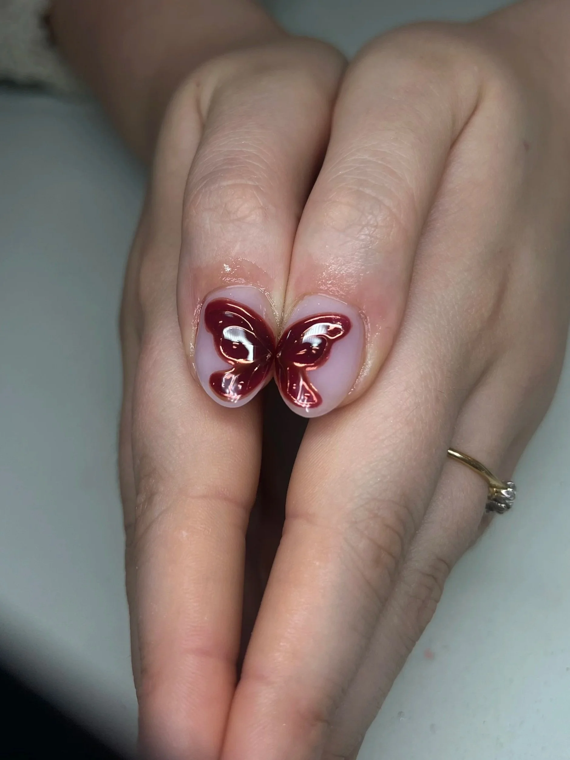 Close-up of a hand with a butterfly design on the thumbnail, featuring brown and white colors, with the person wearing a gold ring on the ring finger.