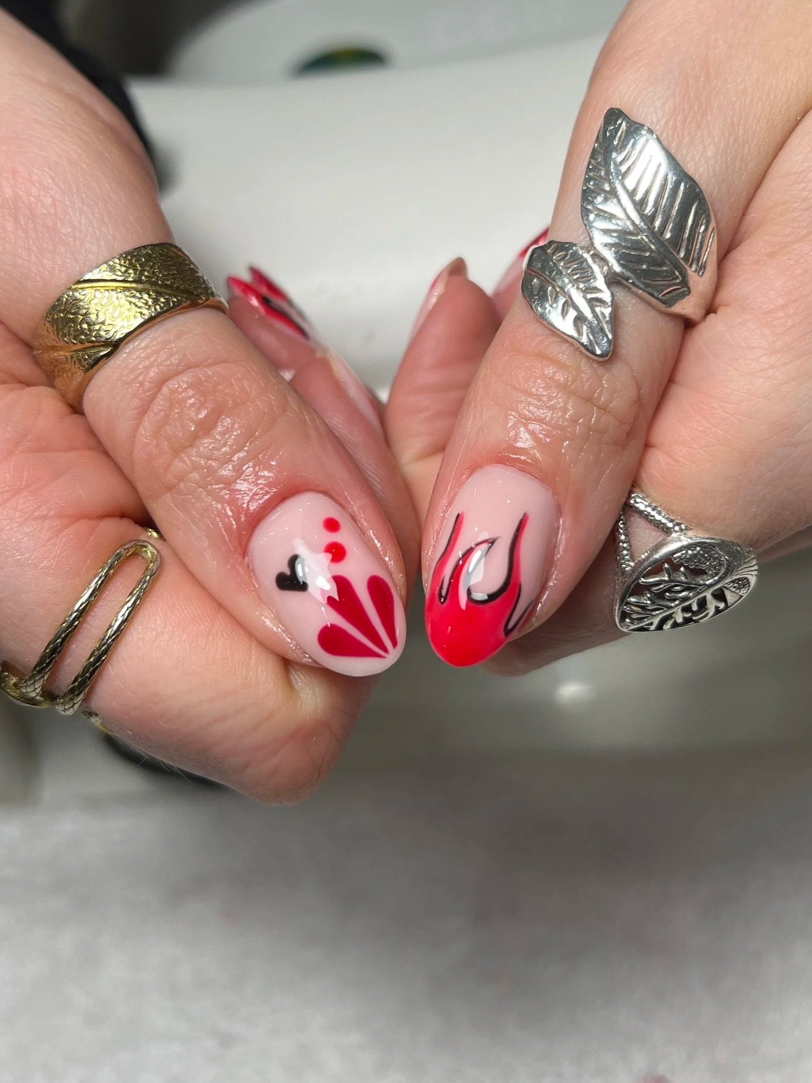 Close-up of a hand with manicured nails featuring red, black, and white abstract designs. The person is wearing three rings, including a silver leaf-shaped ring on the middle finger, a gold textured leaf-shaped ring on the index finger, and a silver 