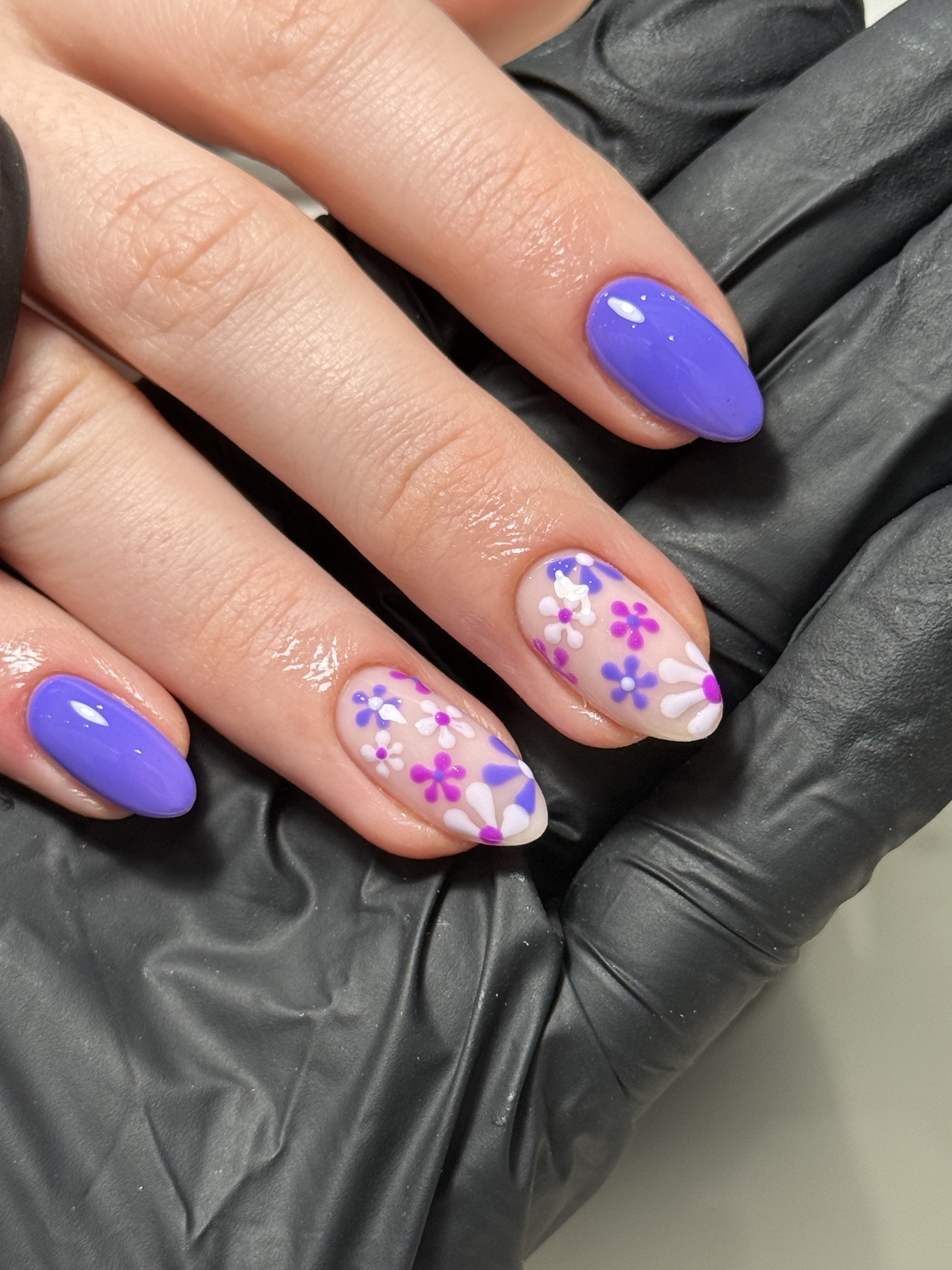 Close-up of a hand with decorated fingernails, showing purple nail polish on some nails and floral nail art on others. The hand rests on a black textured surface.