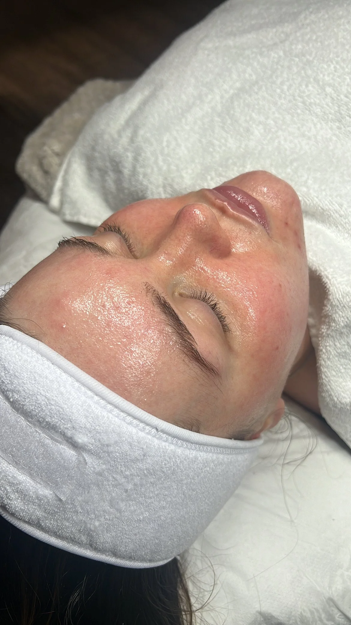 A person lying down with eyes closed, receiving a facial treatment. The person is wearing a white headband and appears relaxed.