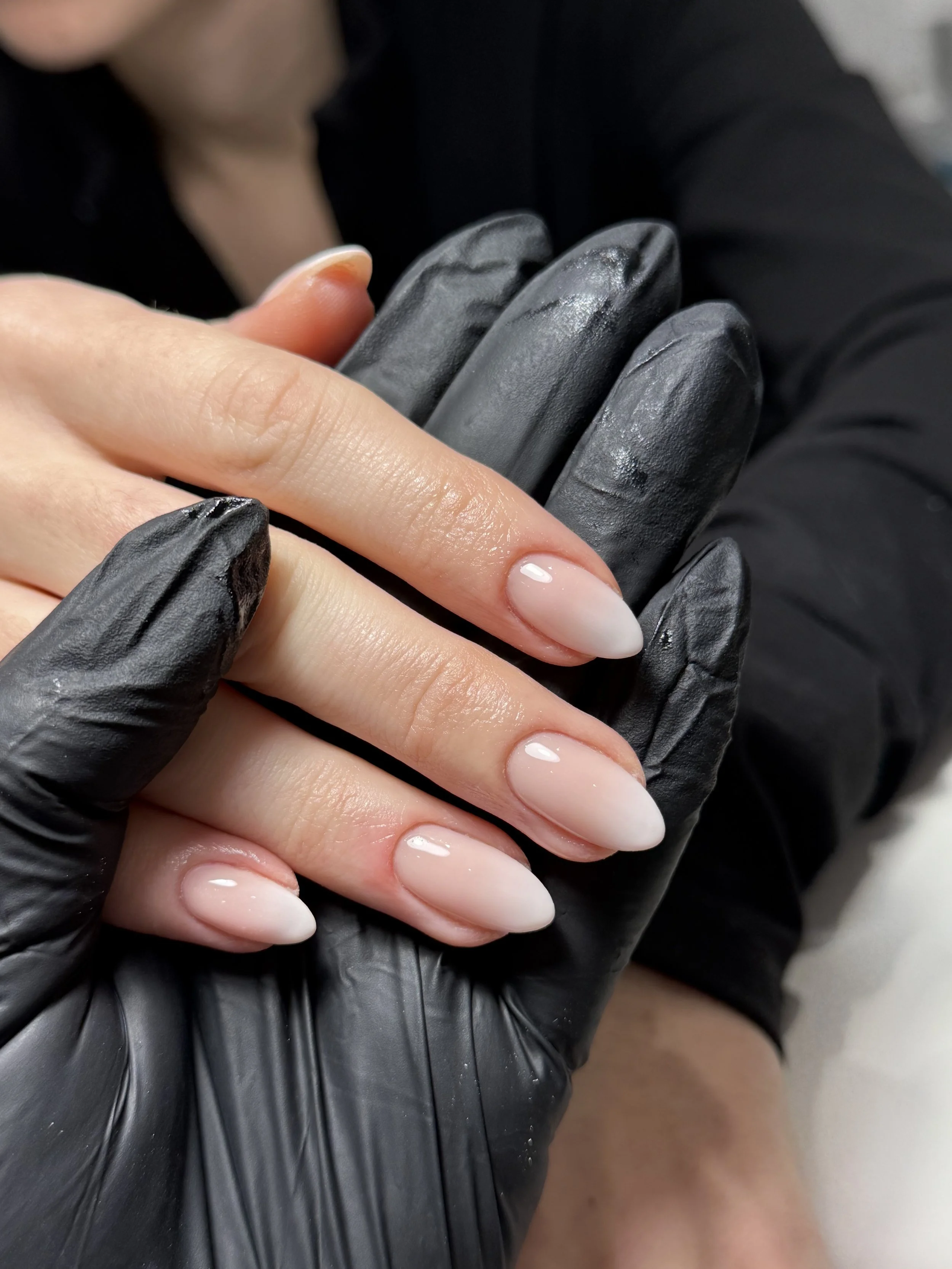 Close-up of a person receiving a manicure, showing the person's fingers with freshly polished, shiny French tips and the manicurist's gloved hand holding the person's hand.