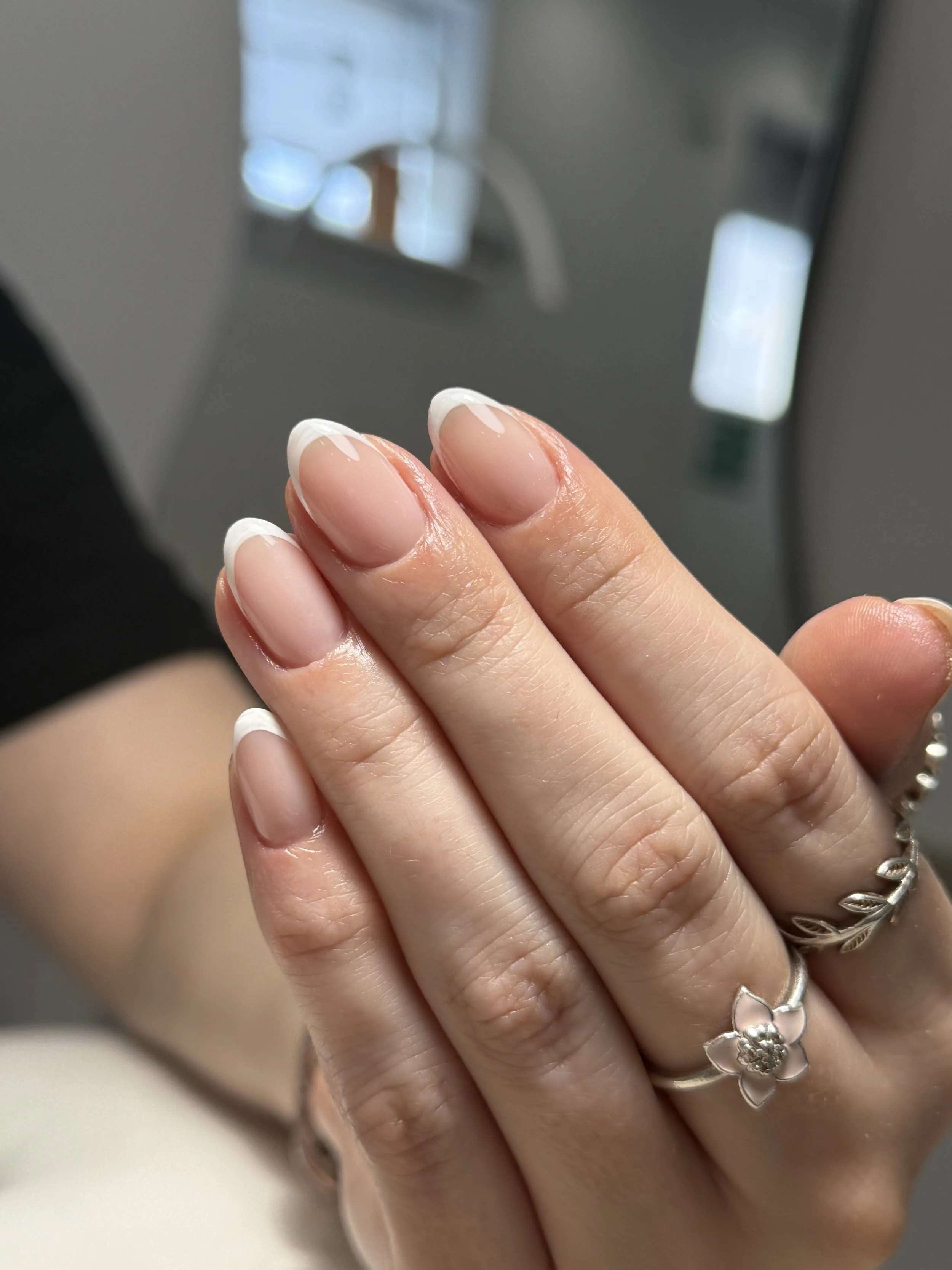 Close-up of a hand with manicured nails and three silver rings, including one with a flower design. The nails are natural with a clear glossy finish and white tips.