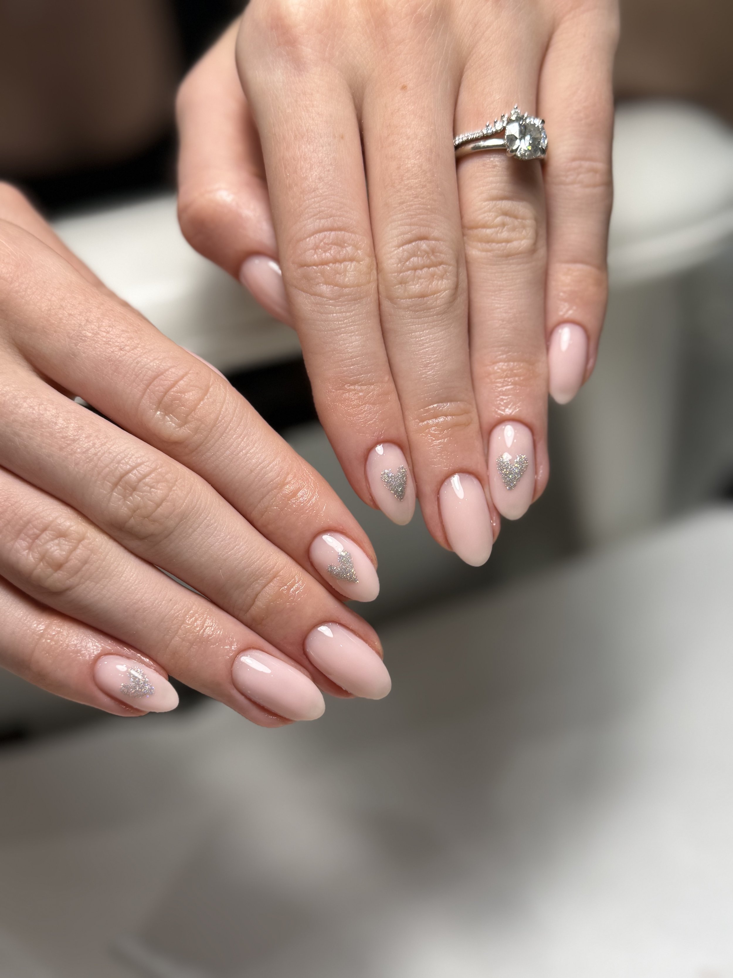 Close-up of a woman's hand showing an engagement ring with a large central diamond and smaller diamonds along the band. The woman's nails are manicured with light pink polish and silver glitter hearts.