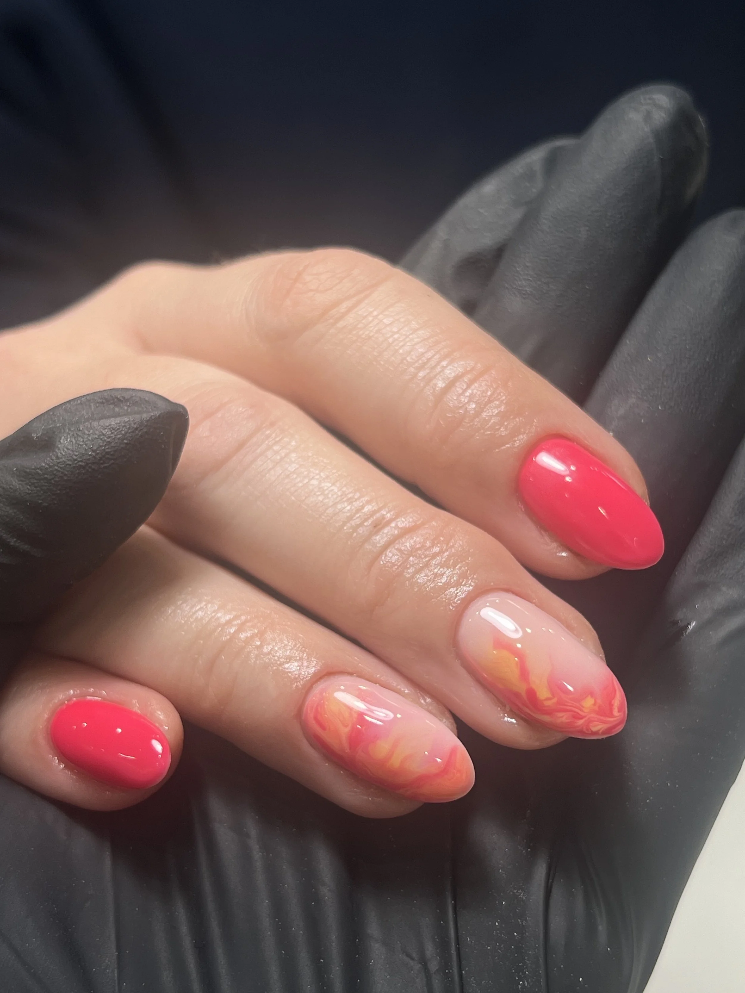 Close-up of a person's hand with bright pink and marbled orange and pink gel nails, with a black glove partially covering the hand, against a dark background.